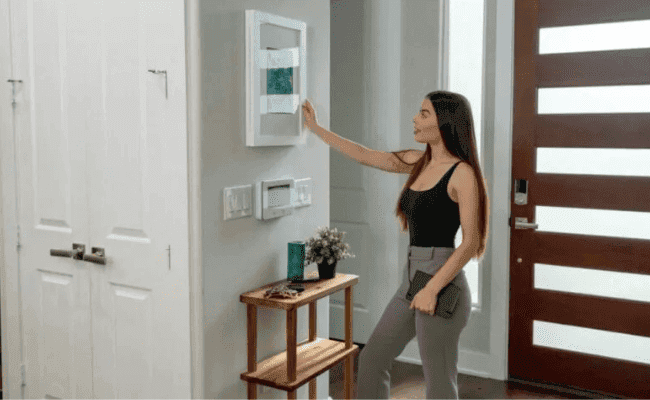 Uma mulher jovem com cabelo escuro e camisa preta sem mangas, em pé perto da porta de entrada de uma casa moderna. Ela está ajustando um painel de controle inteligente na parede, que parece ser um termostato ou um sistema de segurança, com um smart lock visível na porta ao lado. A cena sugere controle e segurança residencial.